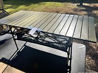 Aluminum camp table with olive green slatted top and black mesh storage shelf underneath, photographed outdoors on a black surface.