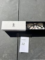 Photo of white SOJOS case, black framed glasses, and tag with lot number 32 on a gray surface