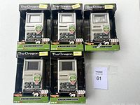 Front view of 5 unopened Oregon Trail handheld video game packages, black and green design with visible screen and buttons