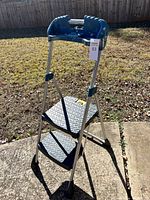 Front view of the folding 2-step stool showing blue handle, metal frame and black steps outdoors.