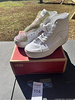 Pair of Vans high top shoes placed on a red Vans shoe box outdoors, showing left shoe covered in rhinestones and right shoe plain white