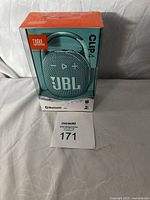 Front view of the JBL Clip4 Bluetooth speaker in box showing the speaker, JBL logo, and Bluetooth and battery icons.
