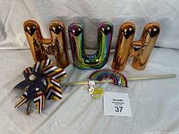 Four large letter figurines spelling out 'H U H', each in shiny metallic finishes with two gold toned and one iridescent. Pinwheel and rainbow decorations arranged in front.