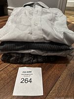 Four folded dress shirts stacked on wooden floor, including striped and patterned shirts, with MaSold lot number card 264.