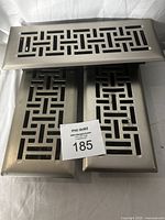 Three metal vent covers, one large and two smaller, arranged on white cloth background. Each vent cover has a maze-like rectangular cut-out pattern.