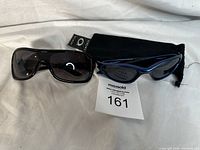 Two pairs of sunglasses including a black Oakley pair with soft pouch and a blue unbranded pair, both laid on a white surface.