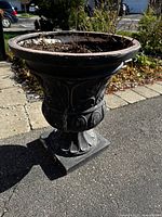 Full view of the black decorative outdoor planter showing its urn shape and floral design, placed outside on pavement.