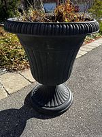 Front side view of black plastic urn planter with ribbed details and decorative rim, filled with soil and dried plants.
