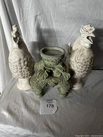 Front view of two off-white ceramic rooster jugs flanking a central green elephant-shaped candle holder with intricate carvings and a round holder on top.