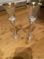 Pair of clear glass candleholders with gold filigree at the rim displayed on wooden floor.