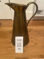 Front view of the tall hammered brass pitcher showing its flared spout and curved handle on wooden flooring.