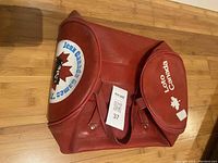 Red leather square bag with front flaps showing 'Loto Canada' and 'Jeux Canada Games 1977' logos on a wood floor background.