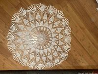 Top view of round hand crocheted white lace doily/tablecloth laid flat on wooden floor, showing full pattern and scalloped edge.