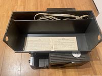 Top view of the Vintage Sawyers 550A projector with the power cord stored inside the open compartment and the instruction label visible inside.
