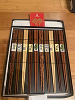 Set of 10 wooden chopsticks in a black box with white interior, showing various wood grain patterns and Japanese labels on each pair, neatly arranged side by side.
