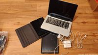 Overview of MacBook Pro open next to two iPads, cases, and power adapters on hardwood floor