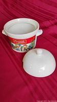 White ceramic Campbell's Soup soup tureen with lid removed, placed on red fabric background. Shows side handles and colorful Campbell's label with child figures.