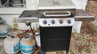 Front and side view of the Pit Boss gas grill showing the four burner knobs and side burner knob with four propane tanks arranged beside it on ground. Some tanks show rust and wear.