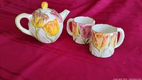 Photo of hand painted teapot and two matching teacups with tulip floral motif on a red cloth background.