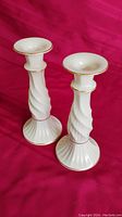 Two tall white porcelain candlesticks with elegant spiral textured design and gold trim detail on red fabric background.