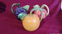 Three vintage Majolica pitchers shaped as grape bunch, orange, and peach, arranged on red cloth background.