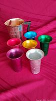Photo showing aluminum pitcher with black handle and six anodized colored wide rim cups arranged on red fabric background.