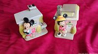 Photo showing two Disney ceramic canisters shaped like houses with Mickey and Minnie Mouse figures attached, placed on a red surface