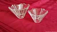 Two thick, clear crystal dishes with angular, geometric shapes viewed from a side angle on a red fabric.