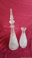 Pair of two glass decanters on a red background, one with stopper and starburst pattern, the other smaller with floral texture.