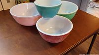 Four Pyrex mixing bowls in pink, teal, green and red, arranged on a table showing all colors and sizes
