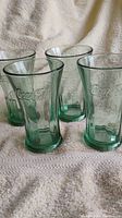 Four heavy green Coca-Cola wide rim glasses shown upright on a white textured fabric surface, highlighting embossed logos and flared rims.