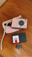Pink Sony Cyber-shot digital camera with battery, charger and memory card on wooden surface, pink protective pouch in background.
