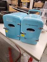 Photo of front and side angles of two light blue mini Frigidaire refrigerators on table showing intact doors with oval windows and silver handles.