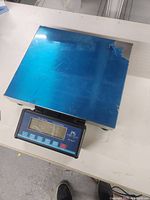 Top-down view of stainless steel weighing platform with a blue protective film partially peeled off and digital display unit beneath it.