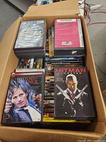 Overhead photo of a cardboard box filled with multiple DVDs including visible titles like A History of Violence and Hitman Unrated.