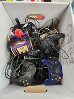 Top view of a box containing multiple tangled game controllers including Xbox controllers, a yellow Wii Party controller, and a red ball joystick.