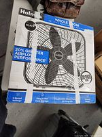 Close-up of front of boxed Holmes fan showing branding and features including 20% greater airflow, 3 speed settings, durable frame, easy cord storage, black color, 20-inch blade, taped shut.