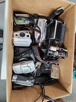 Top-down view of a box containing multiple vintage cameras and camera accessories, including a white compact camera and a black camera with strap.