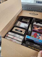 Cardboard box with several music cassette tapes seen from above and one side, showing various album covers and labels.