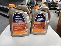 Two beige plastic jugs of PEAK antifreeze and coolant with orange caps on a table. One jug appears full and sealed, the other half full.