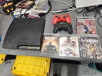Photo showing Sony Playstation 3 console, two controllers (one red third-party, one black Sony), and four PS3 games arranged on table.