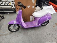 Side view of a purple electric moped with a white seat and floral decals placed on concrete floor indoors.