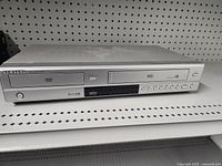 Front view of silver Samsung VCR DVD combo unit showing disc tray, VHS slot, power button, and control buttons on shelf.