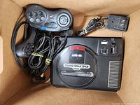 Top view of Sega Genesis 16-BIT console with wired controllers and power supply inside a cardboard box, shows console label and power adapter.