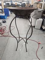 Full view of the standing metal brazier with wide bowl and curved wrought iron legs in a workshop environment.