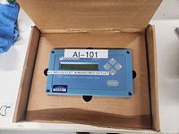 Blue rectangular fluid measurement instrument with label AI-101 in open cardboard box.