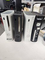 Four Xbox 360 consoles standing upright, showing front sides with power buttons and disc trays. One black console lacks a front faceplate.