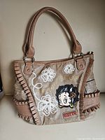 Front view of taupe colored Betty Boop handbag showing embroidered flowers, gold lettering, Betty Boop face applique, and ruffle trim details on faux leather material.