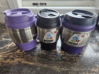 Photo showing three insulated Bubba mugs on a countertop: two larger 1.5 litre mugs with purple and black lids, and one smaller 1 litre mug with a black lid. All have stainless steel bodies and plastic bases.