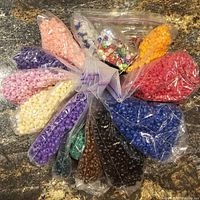 Multiple plastic bags arranged in a circle on a countertop showing a variety of colored craft beads including seed beads and shaped beads.
